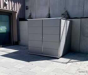 Ein moderner, grauer Gartenschrank steht auf einer Terrasse neben einem Gebäude mit Fenstern und Pflanzen.