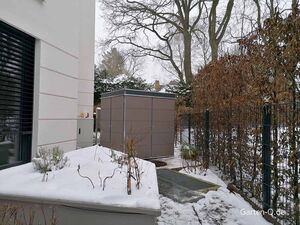 Ein verschneiter Garten mit Zaun, kahlen Bäumen, einem Schuppen und einem modernen Gebäude.