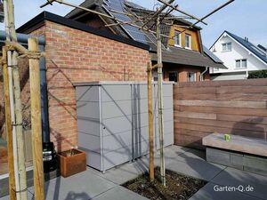 Ein moderner Garten mit grauem Schrank, Holzzaun, Bank und Bäumen. Im Hintergrund ein Haus mit Solarpanels.