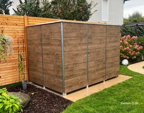 Ein Holz-Gartenschrank steht neben einem Holzzaun in einem Garten mit Pflanzen und Rasen.