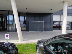 Ein moderner Gebäudeeingang mit Glasfront, zwei schrägen Säulen und einem Halteverbotsschild. Autos parken davor.
