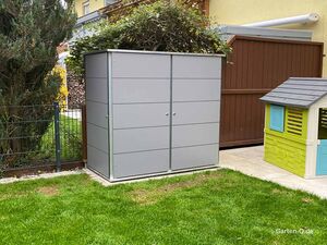 Grau Metallschuppen, Kinderspielhaus, grünes Gras, neben Zaun und braunem Holzschuppen.