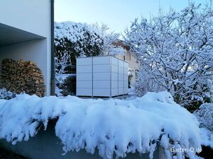 Ein verschneiter Garten mit Holzstapel und einem weißen Gartenhaus im Winter.