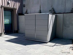 Ein moderner, grauer Gartenschrank steht auf einer Terrasse neben einem Gebäude mit Fenstern und Pflanzen.