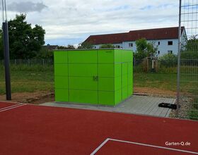Ein grüner Schuppen auf einer Betonfläche neben einem roten Sportplatz, umgeben von Zaun und Bäumen.