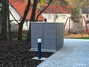 Ein grauer Müllcontainer oder Schuppen neben einem gepflasterten Weg, mit Bäumen und Schaukel im Hintergrund.