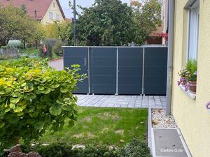 Ein Paketkasten kombiniert mit Mülltonnenboxen im Vorgarten neben einem Haus mit Blumen auf der Fensterbank und umgeben von Bäumen und Rasen.