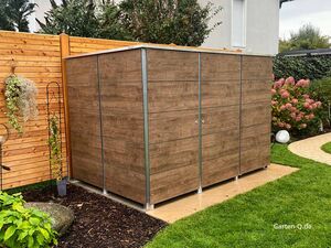 Ein Gartenhaus aus Holzplatten steht in einem gepflegten Garten neben einem Holzzaun und Blumen.