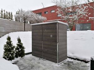 Ein graues Gartenhaus im Schnee, umgeben von schneebedeckten Bäumen und einem roten Backsteingebäude im Hintergrund.